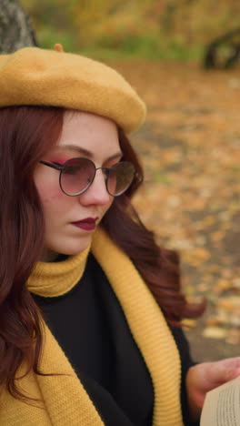 dama con labios pintados con boina amarilla sentada al aire libre, descansando en un árbol, hojeando un libro mientras coloca la mano en una página en particular, hojas de otoño esparcidas por el suelo