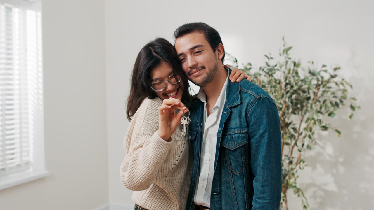 Young couple in love renting buying their first apartment together joyful woman embraces boyfriend in hand holding keys to flat they stand in the middle of a newly renovated empty room