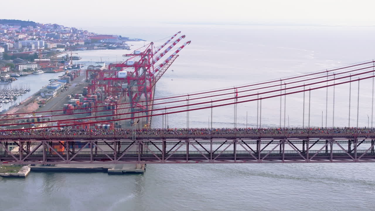 Aerial side-on drone view of thousands of half marathon and 10k runners crossing the red 25th April suspension bridge beside the Lisbon waterfront, marina, port docks, industrial cranes in Portugal