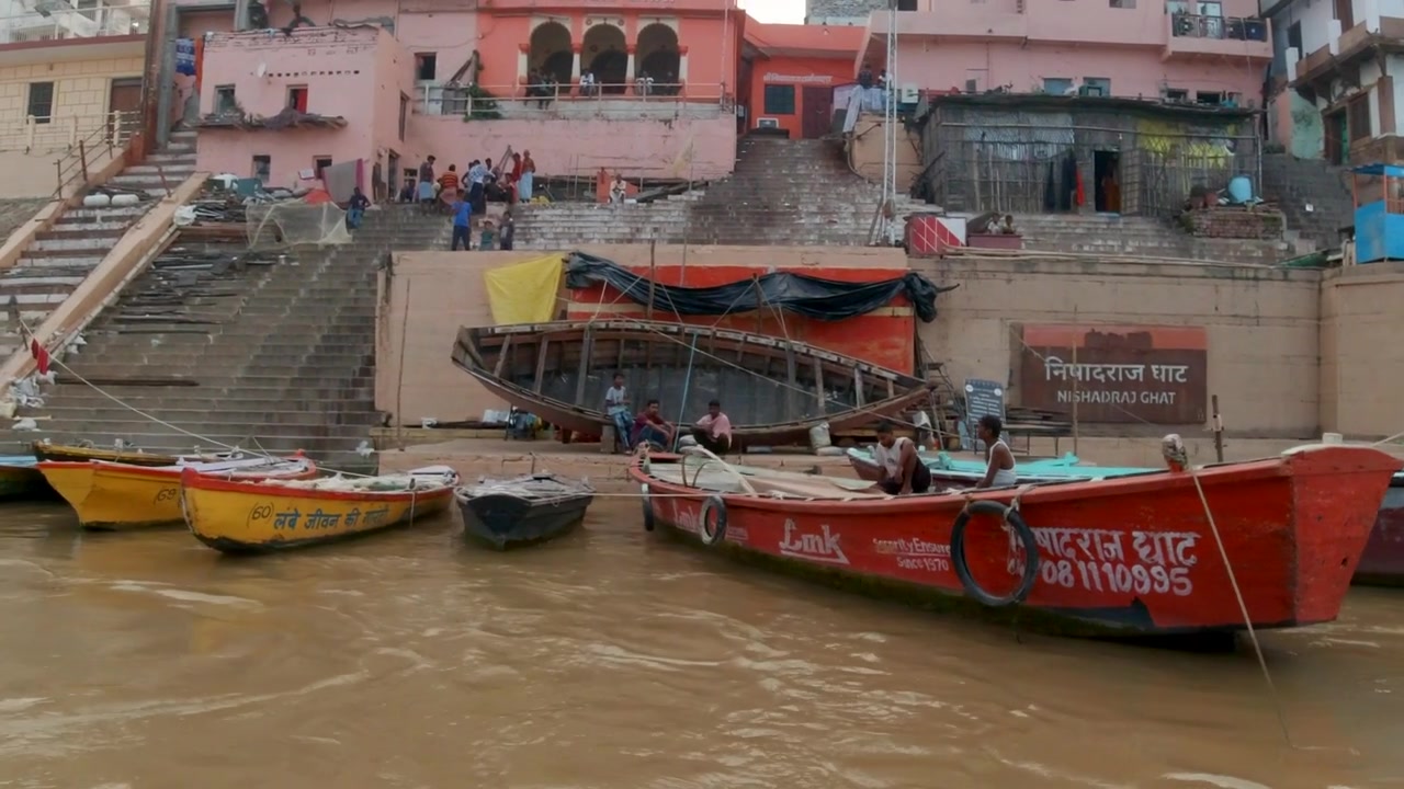 Sinematik Varanasi Hindistan Ganj Nehri tekne turu kanal tekneleri Kuzey Eyaleti kıyıda büyük kalabalık Antik Kutsal şehir Ghat Pradeş Eyaleti manzarası gri bulutlu öğleden sonra çamurlu kahverengi sola doğru