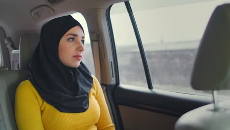 young muslim woman in hijab sitting in car on passenger rear seat. muslim woman day dreaming in back seat. a young woman in a hijab looks out the window.