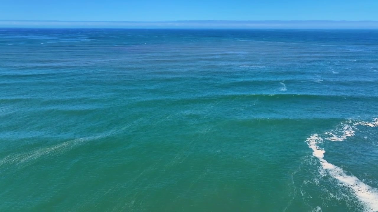 Riprese aeree mostrano le onde dell'Oceano Atlantico che si infrangono lungo la spiaggia sabbiosa. La costa presenta dune costiere e distese di acqua blu sotto cieli estivi sereni