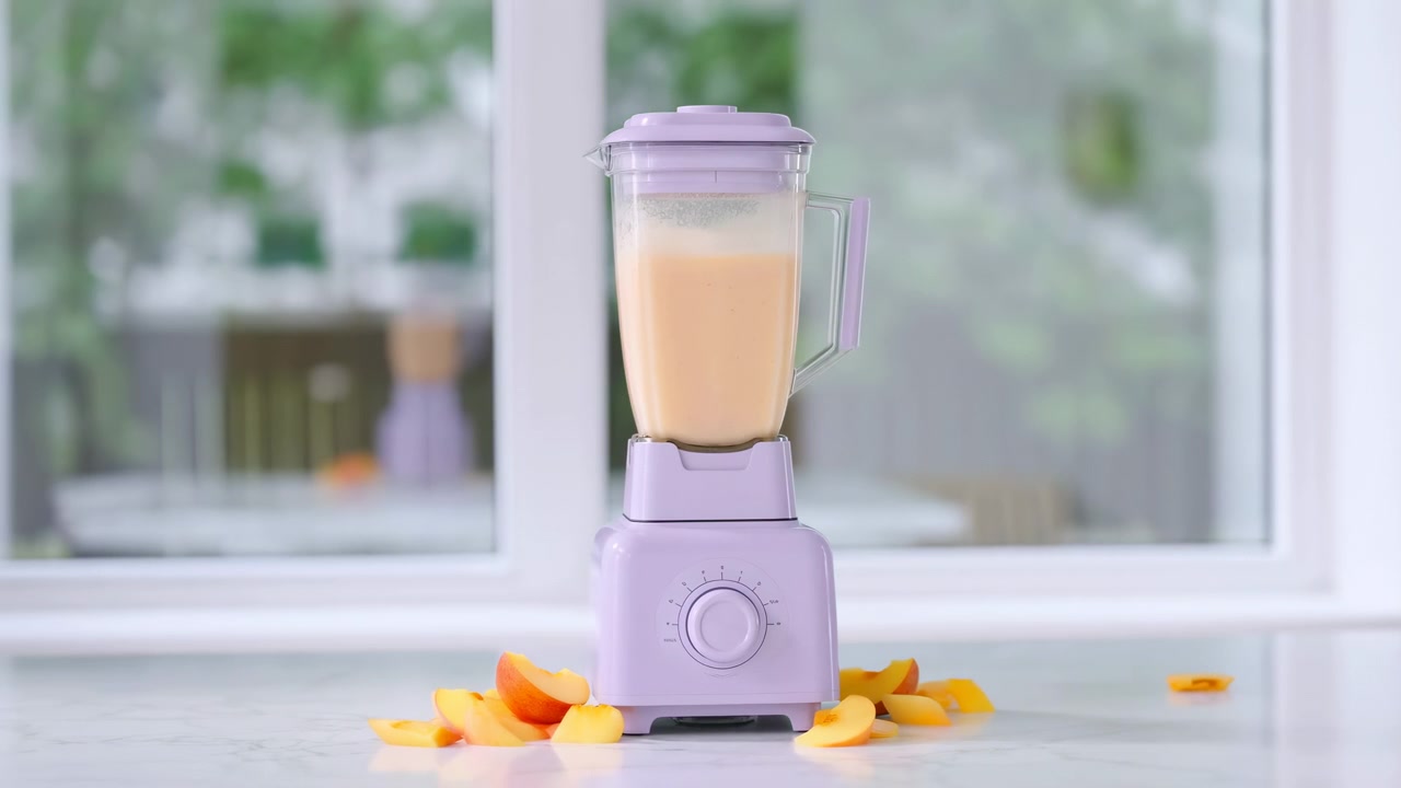 En lavendelfarvet bordblender fyldt med en cremet ferskenfarvet smoothie står på en ren køkkenoverflade, omgivet af skåret frugt og et par friske.