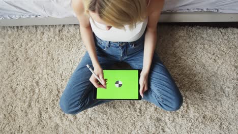 mujer en vaqueros se sienta en posición de loto en el suelo y utiliza tableta electrónica con pantalla verde, vista superior. maqueta. mujer talentosa dibuja con lápiz en la tableta. concepto de aparatos digitales y creatividad