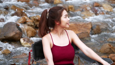 Asian-woman-in-red-sitting-on-a-chair-with-big-rocks-and-water-slow-splashing-for-relaxing-and-happiness-in-the-vacation-summertime