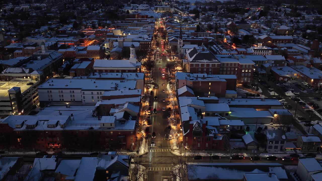 Vista aérea con dron de una pequeña ciudad estadounidense de noche durante una nevada invernal, con farolas brillantes, tráfico a lo largo de la calle principal y edificios cubiertos de nieve. Ambiente cálido y festivo de vacaciones en EE. UU. Tierra.