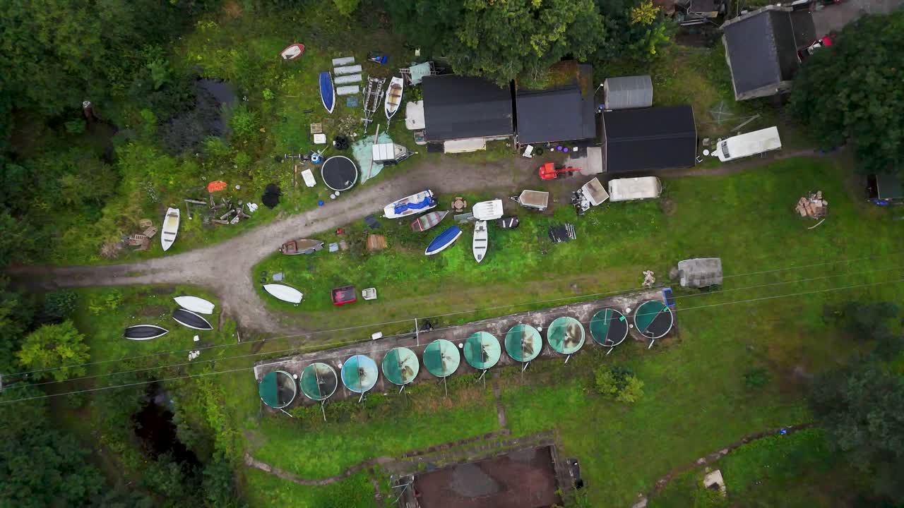 Drone camera smoothly ascends above a cluster of boathouses, small boats, and circular tanks in a green, rural Hope Valley landscape under diffuse daylight