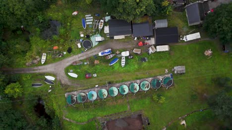 drone camera smoothly ascends above a cluster of boathouses, small boats, and circular tanks in a green, rural hope valley landscape under diffuse daylight