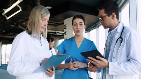 Caucasian-male-and-female-doctors-speaking-with-young-nurse-while-typing-on-tablet-screen-and-writing-information