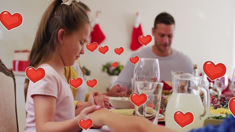 Animation-of-hearts-floating-over-happy-caucasian-family-during-dinner-praying-at-christmas