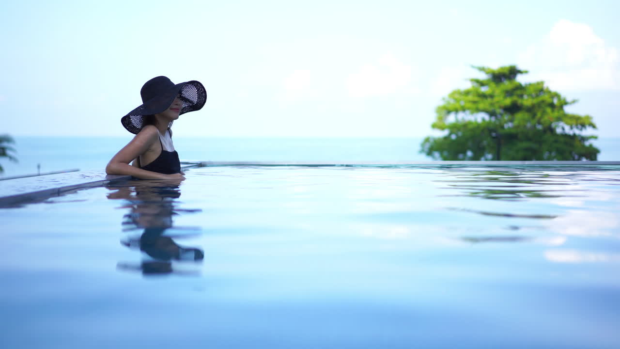 Elegante Dame mit schwarzem Sonnenhut lehnt friedlich am Rand eines entspannenden Infinity-Pools, während das kristallklare Wasser mit ihrem Spiegelbild leichte Wellen schlägt.