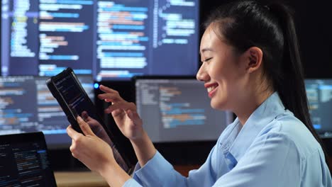 close up side view of asian female programmer looking at database on tablet while writing code by a laptop using multiple monitors showing database on desktops in the office