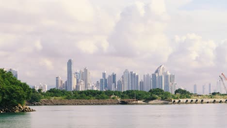 handheld shot of landscape with skycraper skyline in panama city's bay waterline viewed from amador's causeway during a sunny summer day behind dense vegetation mangrove and a cloudy blue sky