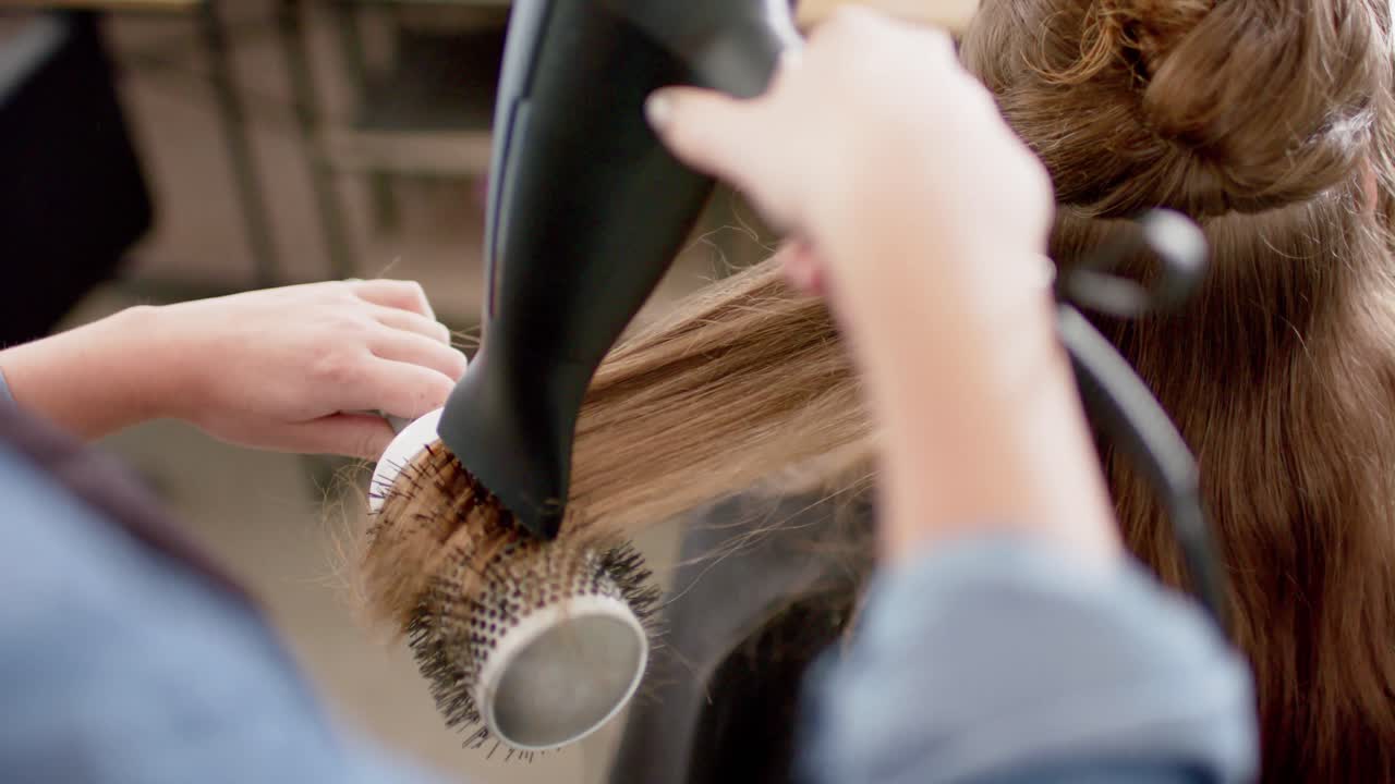 Caucasian female hairdresser styling client's long hair with hairdryer and brush, in slow motion