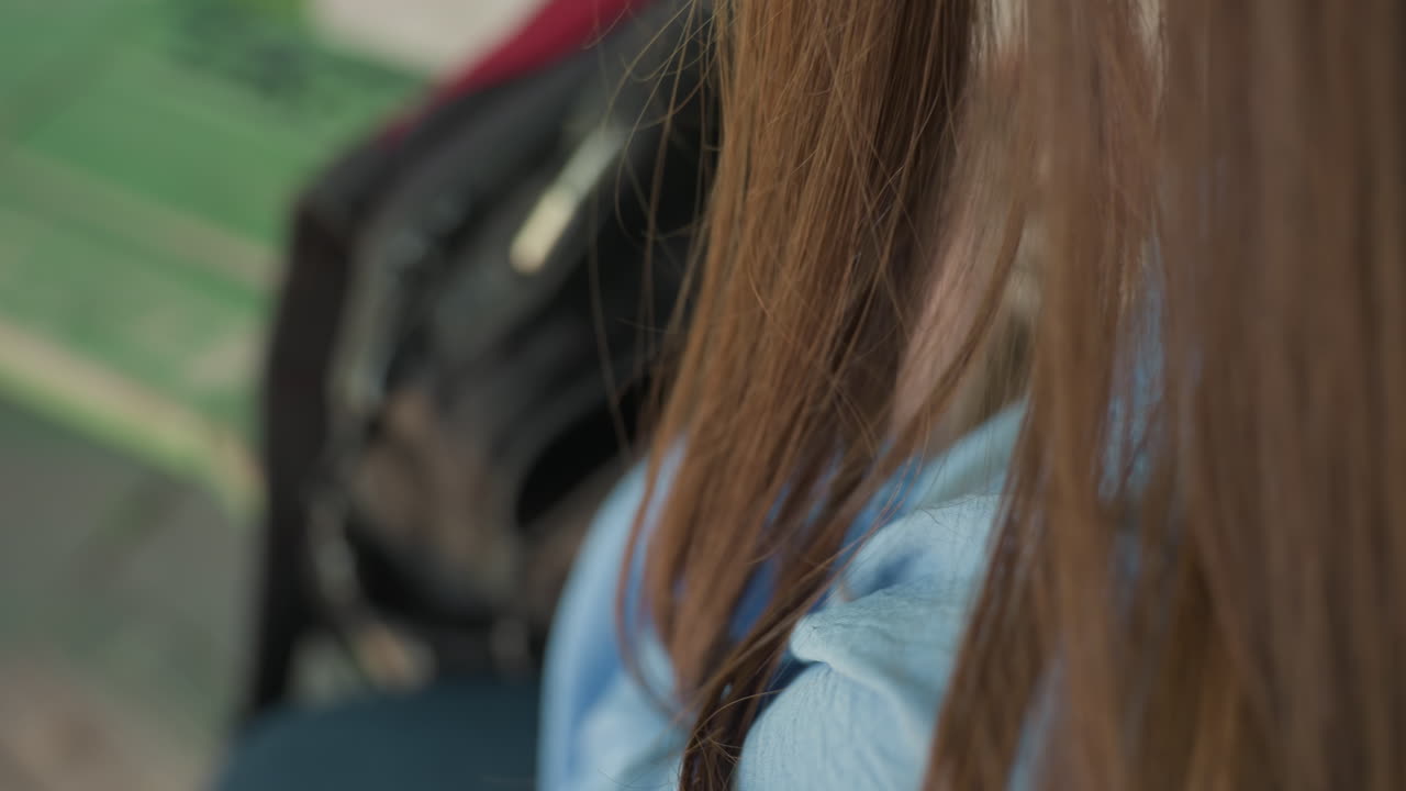 젊은 백인 여성이 벤치에서 배낭을 찾고 있습니다. 파란색 셔츠를 입고 긴 머리가 어깨 위로 늘어져 있으며, 지퍼 포켓을 샅샅이 뒤지는 손이 클로즈업되었습니다. 도시 공원 좌석, 아침 햇살, 집중된 분위기입니다.