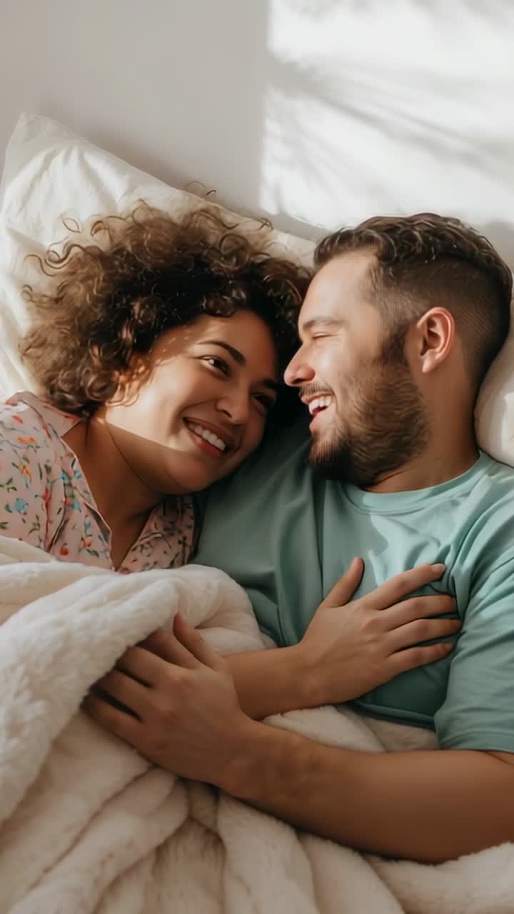Vertical video: Sun waking couple, woman in floral pjs, smiling, touching chest on bed with blanket