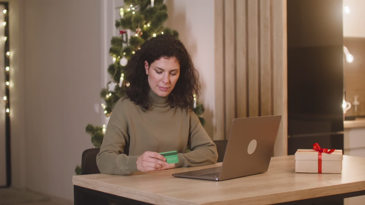 mujer morena comprando en línea con una tarjeta de crédito usando una computadora portátil sentada en una mesa cerca de un regalo en una habitación decorada con un árbol de navidad 1