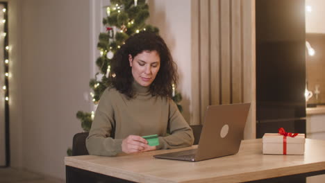 brunette woman buying online with a credit card using a laptop sitting at a table near a present in a room decorated with a christmas tree 1