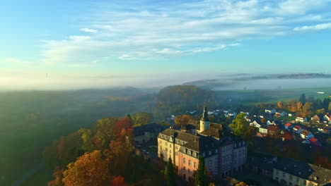 Vista-Aérea-Del-Castillo-Y-El-Pueblo-En-El-Pintoresco-Paisaje-De-Alemania-En-La-Soleada-Y-Brumosa-Mañana-De-Otoño,-Disparo-De-Drones