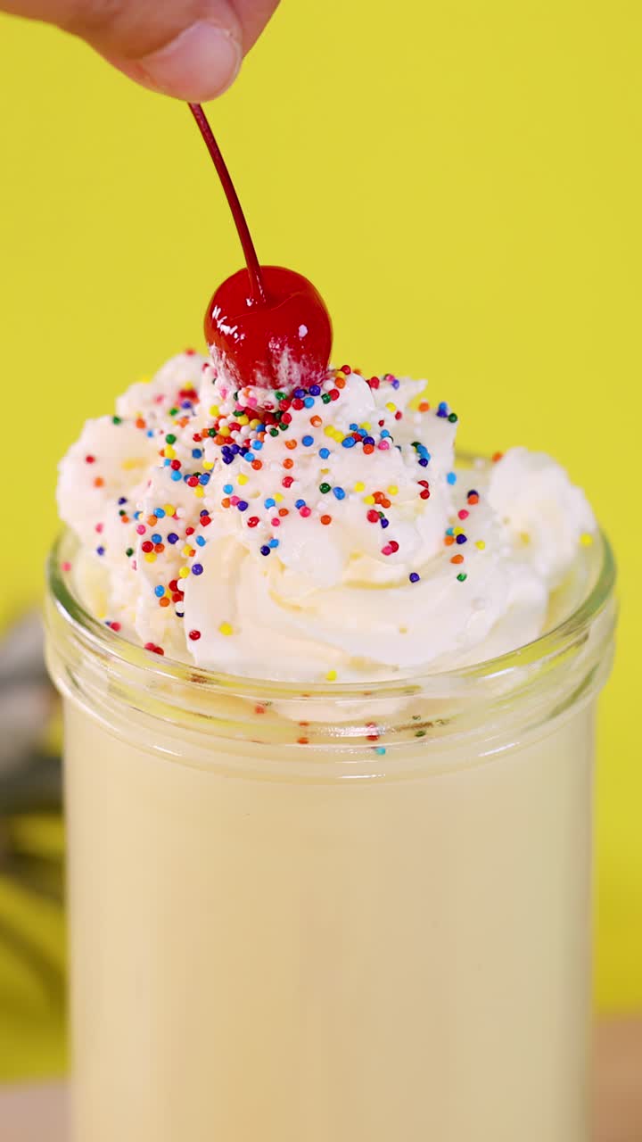 Primo piano di una mano che posiziona una ciliegia rossa su un frullato all'ananas con panna montata su uno sfondo giallo brillante