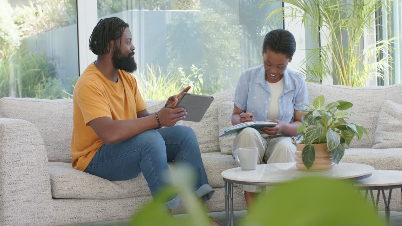 African American pair working on sofa, man raising finger showing tablet, woman writing in notebook