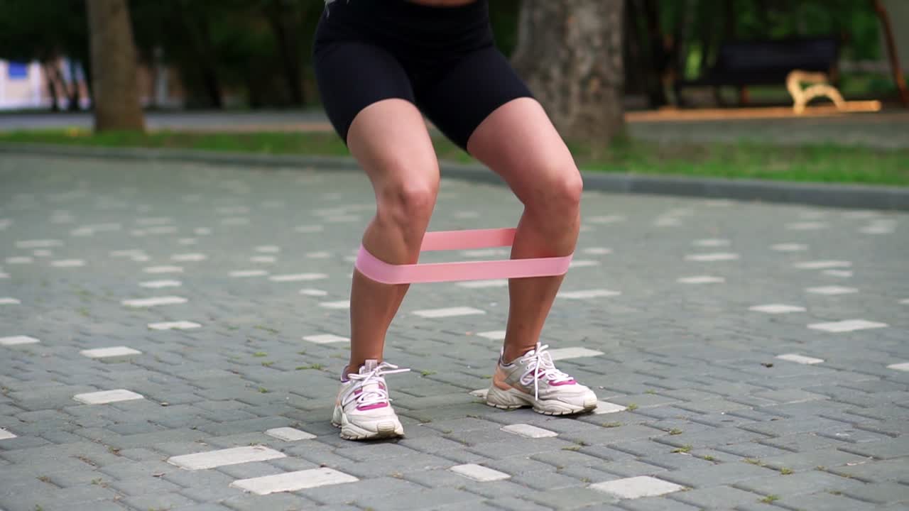 mujer atleta joven irreconocible ejercicios con banda de resistencia rosa haciendo sentadillas al aire libre en el parque público local, verde