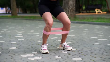 mujer atleta joven irreconocible ejercicios con banda de resistencia rosa haciendo sentadillas al aire libre en el parque público local, verde