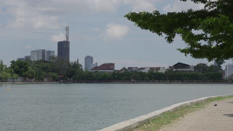 city skyline viewed across a tranquil lake with greenery, under a clear sky, daytime, urban peacefulness