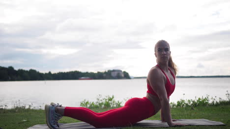 young woman in red sportswear sitting on yoga mat outside doing exercises and practicing yoga