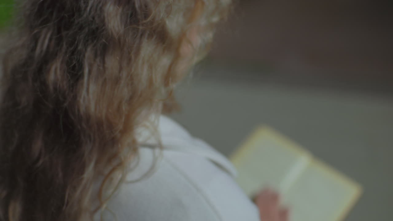Close up rear view of woman seated outdoors at night opening to new page of book in her hand as her curly hair sways gently in wind, slightly blurred for soft and tranquil visual atmosphere