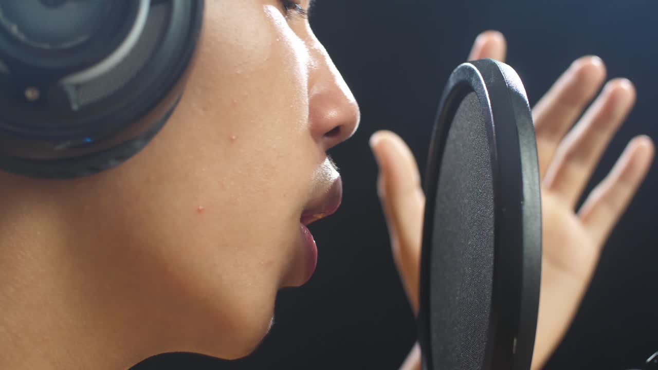 젊은 아시아 소년의 입이 검은 바탕에 있는 콘덴서 마이크로 노래하는 근접한 측면 시각