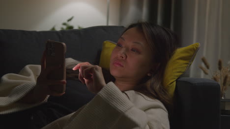 close up of woman spending evening at home lying on sofa with mobile phone scrolling through internet or social media