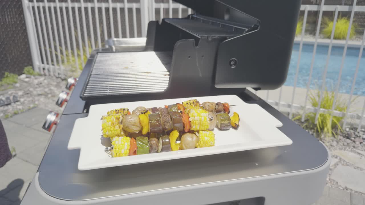 The last vegetable skewer with corn, pepper, zucchini, potatoes and mushrooms is removed from a stainless steel bbq. Side profile of camera following the tongs from the grill to the plate