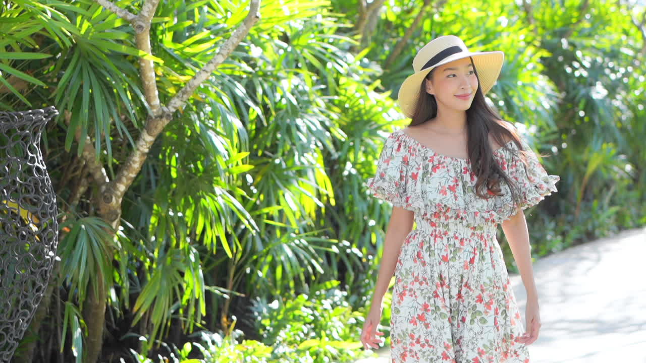 Attractive young Asian woman in colorful floral print sundress with wide brim straw hat walking on path beside tropical plants and deep green island foliage of luxury resort destination