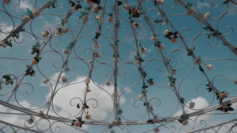 close up shot of a ceremonial metallic arch, decorated with white and pink roses, over a beautiful summer blue sky and white clouds