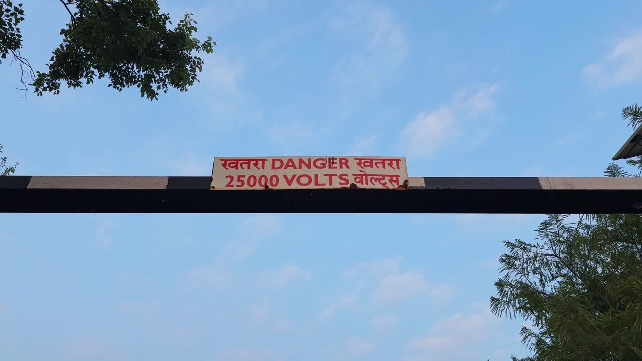 Static shot of a railway barrier displaying a danger sign above the tracks, with bold voltage text set against a clear sky, while surrounding trees frame the elevated guard structure from below