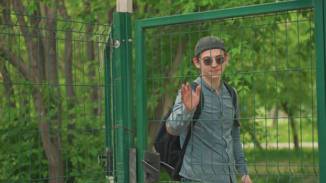 Un hombre abre la puerta de un parque con una mochila, lleva gafas de sol y gorra, toca el pestillo y saluda a su acompañante mientras entra en un camino frondoso detrás de una valla verde, paseo veraniego relajado y natural, naturaleza urbana