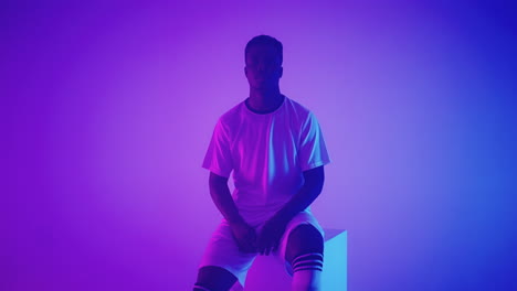 slow motion portrait of a black professional football player in a white uniform looking into the camera in the blue-red neon light of the studio. a brutal football player confident and victorious