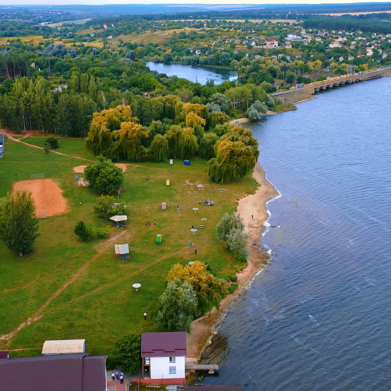Green wooded bank of the river locating in the city. Hotel with the pier and few boats on the water. Top view