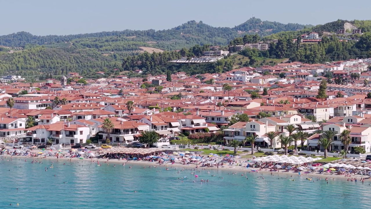Spiaggia affollata di Pefkochori in estate, villaggio di Pefkochori con paesaggio montano e forestale sullo sfondo, Calcidica, Mar Egeo, penisola di Kassandra, ripresa aerea con drone.