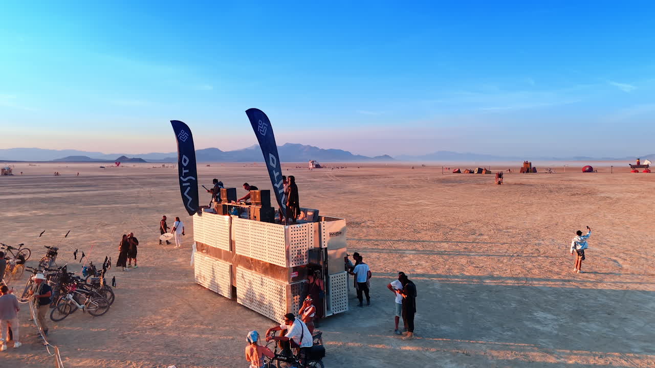 Sunset platform gathering with cyclists. Festival guests circle a multi‑level platform with flags as the sun sets over the distant ranges
