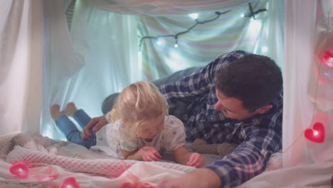 father and young daughter reading story in homemade camp in child's bedroom at home - shot in slow motion