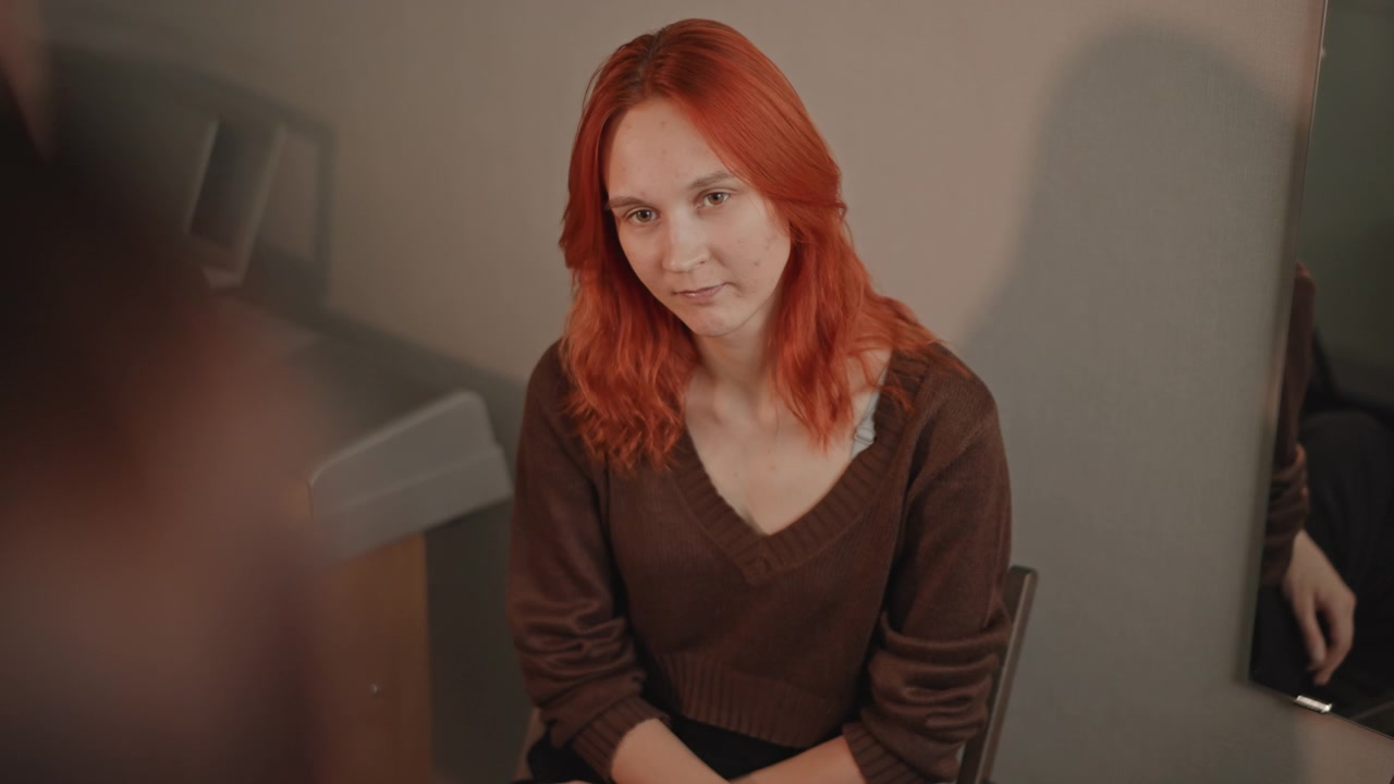 Candidate shows confident composure. Tressed performer awaits assessment with poise and grace. Redhaired artist stands composed as mentor provides gentle feedback during final evaluation