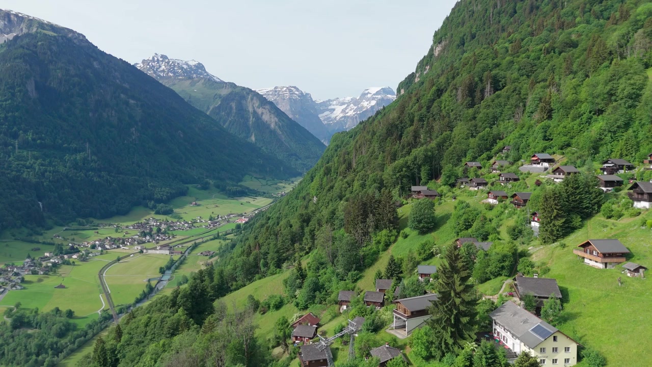 Ampia valle alpina con fiume serpeggiante e villaggio si estende tra cime innevate accanto a chalet in legno su un ripido pendio boscoso nel Canton Glarona, Svizzera. Ampia ripresa aerea con drone.