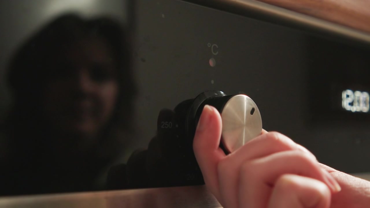Close-up view of hand grasping and turning metallic oven control knob on stainless steel appliance, with blurred face reflected in dark glass door, subtle fingertip pressure, circular rotation, soft