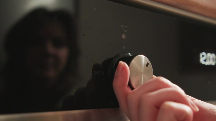 Close-up view of hand grasping and turning metallic oven control knob on stainless steel appliance, with blurred face reflected in dark glass door, subtle fingertip pressure, circular rotation, soft