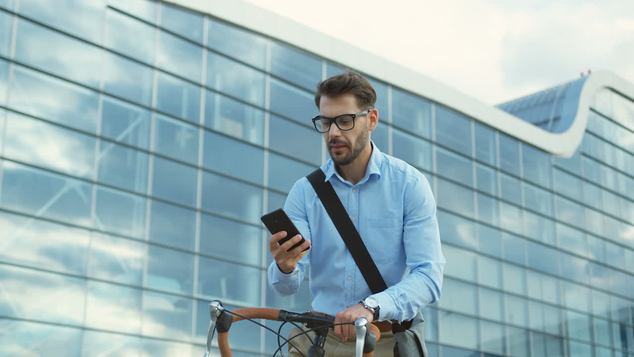 hombre guapo con gafas de pie con una bicicleta, sacando de su bolsillo un teléfono inteligente y comenzando a escribir en él