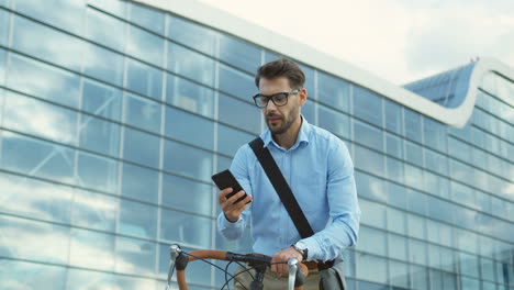 handsome man wearing glasses standing with a bicycle, pulling out of his pocket a smartphone and starting to type on it