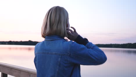 young short haired beautiful woman with jeans jacked fixing her hair and watching pink sunset in front of the lake