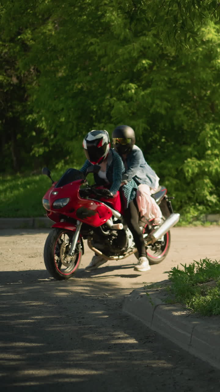 una mujer con una camisa a cuadros sale en una bicicleta eléctrica roja con su amiga sosteniéndola por detrás, viajan por una carretera sombreada con coches pasando, algunos con faros encendidos